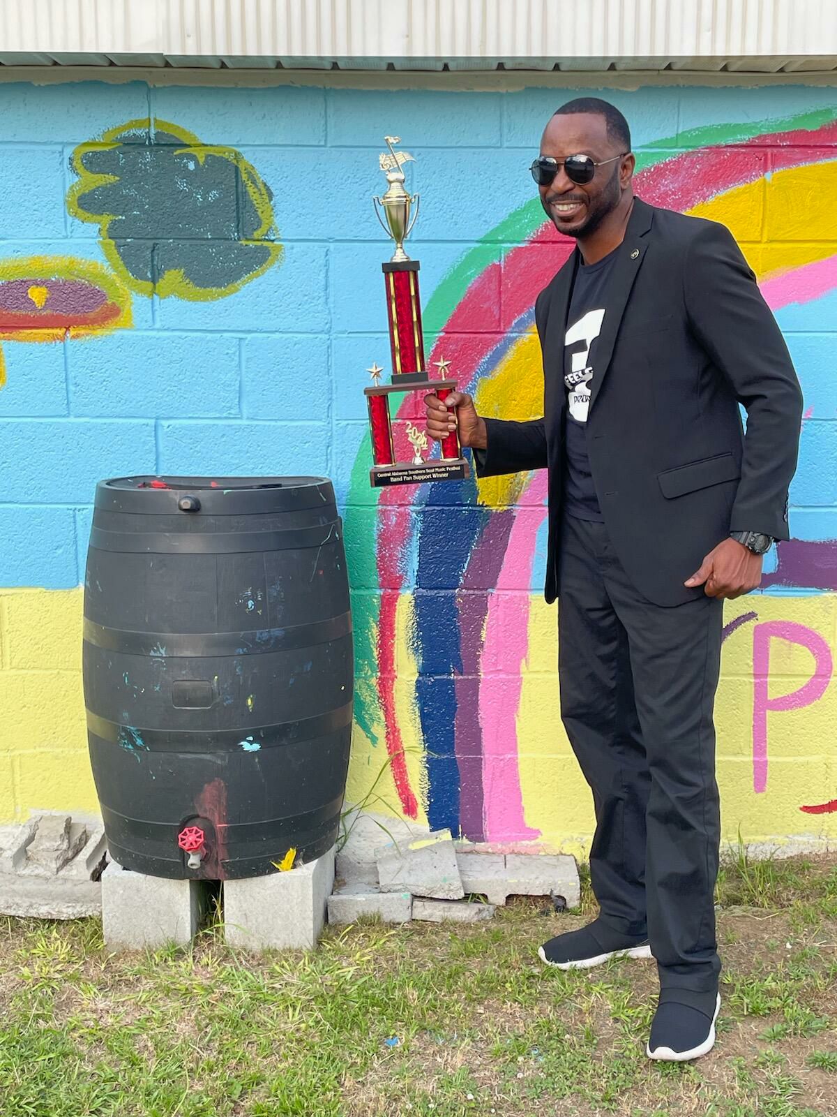 Man in black suit holding a trophy next to a large barrel with a colorful mural in the background.