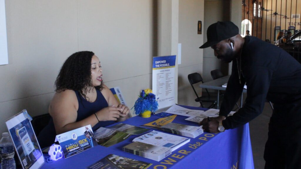 Two people engaged at an informational booth with brochures and a mascot.